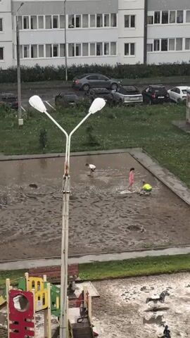 A video shows a playground with several children playing in puddles. In the foreground, there's a street lamp and a portion of a playground structure. In the mid-ground, there's a large, muddy puddle where three children are playing. One child is wearing a white shirt and blue shorts, another is wearing a pink dress, and the third is wearing a yellow shirt and green shorts. They are splashing in the water. In the background, there are several parked cars and a multi-story building. It appears to be raining, as there are visible raindrops in the air and on the ground. In the lower right corner of the image, there is a toy horse and another child is standing nearby.