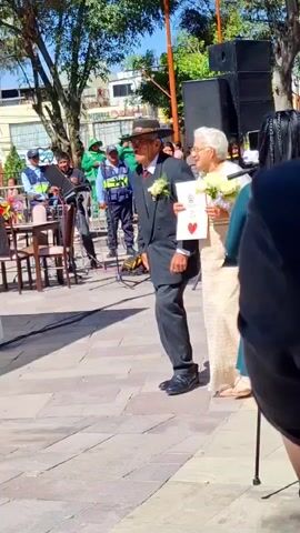 An older couple in formal attire walks together across a brick-paved area. The man wears a dark suit, a hat, and glasses, and the woman wears a cream-colored dress and glasses, carrying a bouquet of yellow roses. The pair appears to be part of an outdoor celebration, possibly a wedding or anniversary. A white drape and some chairs, along with other figures are visible, in the background, suggesting some kind of event space. A photographer holds up a board to block the sun. A few people stand around watching the couple.