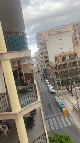 From an elevated position, the video pans across a streetscape. The street is lined with buildings of varying heights and architectural styles, many featuring balconies. Cars and motorcycles are parked along the sides of the street, and some are visible driving. People can be seen walking on the sidewalks below. The sky is overcast, and in the distance, the sea is visible. There are trash bins near a crosswalk.