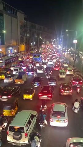 The video shows a high-angle view of heavy traffic at night on a multi-lane road in what appears to be an urban area. Numerous cars, trucks, motorcycles, and auto-rickshaws are visible, mostly stopped or moving very slowly. The scene is brightly lit by streetlights and vehicle headlights. There's a building to the left of the road, which seems to have a pedestrian walkway connected to it. On the right side of the road, a protective barrier separates lanes. Several people are visible on motorcycles, some with passengers. The overall atmosphere indicates a congested urban environment at night. An emergency vehicle siren can be heard in the background.