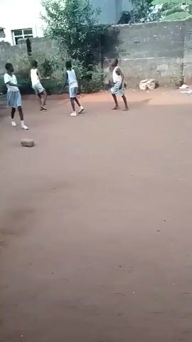 The video shows six children playing soccer in a dirt yard. They are all wearing what appears to be the same white and blue school uniform. The yard is surrounded by buildings and a concrete block wall. There are trees and other vegetation in the background. The children are running around and kicking a soccer ball.
