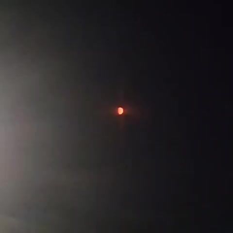 A time-lapse video shows a reddish-orange moon with a sliver of yellow in the center against a dark, hazy sky. The moon appears to be partially obscured by clouds or atmospheric conditions, giving it a diffused glow. The footage shows the moon's position in the sky changing slightly over the short duration of the video, though the overall impression is of stillness. The color of the moon suggests it may be during a lunar eclipse or at moonrise/moonset.