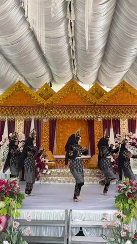 A group of five women in traditional attire are performing a dance on a white stage. They are all wearing black hijabs and elaborate, ornate costumes with gold and black patterns, and distinctive headpieces. The stage is decorated with lush floral arrangements of pink and red flowers on either side. Behind the dancers, a backdrop features a vibrant orange fabric with gold embellishments and elaborate decorative elements with gold trim. The ceiling is draped with white fabric, creating a tent-like effect. The women are moving in unison, with their arms and hands making graceful gestures. Their expressions appear focused and serene.