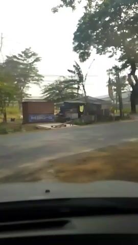 A person wearing a helmet rides a scooter with a box attached to the back. The scooter is on a road with trees and buildings alongside it. The scooter is moving away from the camera. The scene appears to be outdoors in daylight.