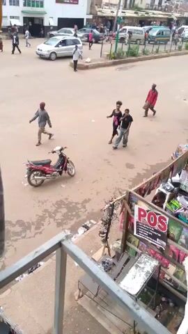 The video shows a street scene with several people and a motorcycle. In the foreground, a red motorcycle is parked. To its right, there is a stall with various items displayed. Further back, two women and two men are walking. One woman is wearing a black dress with pink accents, and the other is in a black t-shirt and jeans. One of the men is wearing a red jacket, and the other is in a black t-shirt and jeans. In the background, there are cars on the street and buildings. The ground is paved with dirt and some puddles.