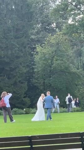 A wedding party is being photographed in a park. A bride and groom are walking away from the camera. A few other people are in the background, also appearing to be guests. The background features trees and a large white building with columns.