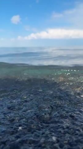 The video shows a low-angle shot of shallow water with a rocky seabed. The water is clear enough to see the pebbles beneath the surface, which are dark and varied in size. Gentle waves lap at the shore, creating ripples and frothy patterns. In the distance, the horizon is visible, where the water meets a bright blue sky dotted with white, fluffy clouds. The overall scene is serene and natural, evoking a sense of peace and tranquility.