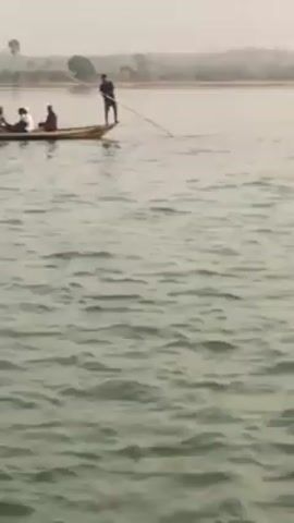 The video presents a series of outdoor scenes on a large body of water, likely a river or lake, consistent with a location like Gburuku, Nigeria.

The initial and concluding frames feature wide shots of the water's surface, showing gentle ripples, with a hazy distant shoreline and subtle vegetation visible under an overcast sky.

Mid-sequence, a young woman with short dark hair, wearing an off-the-shoulder olive-green top and a silver cross pendant, is shown from the chest up. She looks to her right with a thoughtful or contemplative expression, her right hand resting on her chin. Partially visible behind her is a man wearing a patterned headscarf and a white shirt with the text "SAB" discernable.

Another shot reveals a long, narrow boat, characteristic of a dugout canoe or similar vessel, actively traversing the water. It carries multiple passengers and a motorcycle is positioned at the rear of the boat. The overall atmosphere is calm and peaceful, suggesting daily life or travel on the waterway during daytime under hazy weather conditions.