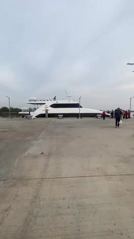 The camera is focused on a large, white speedboat docked at a concrete pier. The boat has black-tinted windows along its side and some equipment on the roof. A handful of people, some wearing red vests, are standing or walking around the boat. One person with a backpack is walking away from the boat, towards the camera. The sky is overcast. The pier has several tall light poles along its edge. In the distance, some trees are visible. The concrete pier surface shows signs of wear and staining.