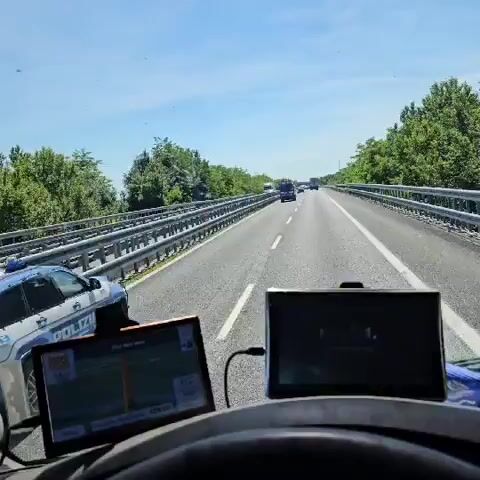 The video shows a view from a driver's perspective on a highway. The driver is following a police car, a blue and white Jeep Grand Cherokee with the Italian license plate PD 4200. The police car is driving in the right lane, with several other vehicles, including trucks, ahead of it. The sky is clear and blue. The video appears to have been taken during the day, in good weather. There are trees visible on the sides of the highway. There are two monitors on the dashboard in front of the driver, one is off and one appears to be a GPS screen.