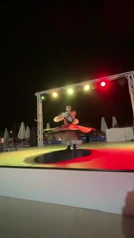 A single male dancer in traditional attire performs a whirling dervish dance on a circular platform stage under color-changing stage lights. The stage is white with a metal frame holding spotlights. The background shows dark sky, beach umbrellas and lounge chairs suggesting an outdoor nighttime performance. The dancer spins rapidly, creating a blur of fabric. The lights shift between purple, red, and yellow.