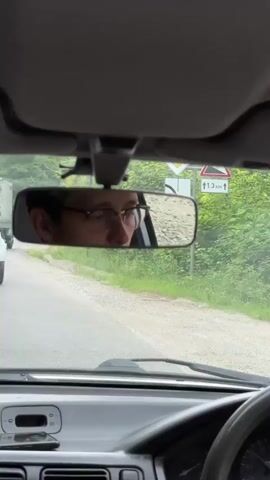 The video shows the interior of a car. The camera is positioned behind the steering wheel, looking forward. The rearview mirror is visible at the top center of the frame, reflecting the face of a person wearing glasses and looking forward. Outside the car, a road is visible with greenery on either side. Another white car is seen in front of the car in which the video is being filmed. The white car is driving away from the camera on a winding road. The dashboard of the car is partially visible at the bottom of the frame. The overall impression is of a person driving on a rural road.