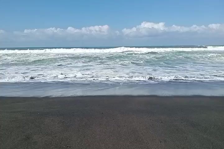 The video shows a wide shot of a beach with black sand in the foreground. The ocean is in the background with waves breaking and white foam washing onto the shore. The sky is blue with scattered white clouds. The scene appears to be calm and peaceful. There are no people or objects visible in the video other than the natural elements of the beach and ocean.
