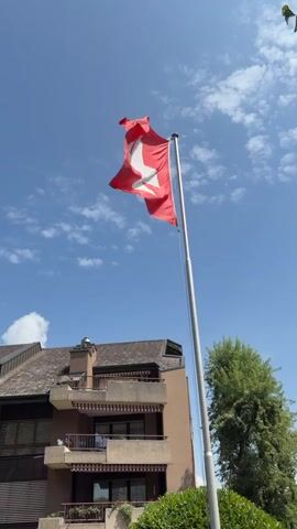 A Swiss flag waves from a flagpole against a clear blue sky with scattered clouds. The flag is red with a white cross. Below the flagpole, a building with balconies and awnings is visible. To the right of the building, a green tree with leaves is present. The sun is shining brightly, casting shadows. The camera is pointed upwards, capturing the flag from a low angle.