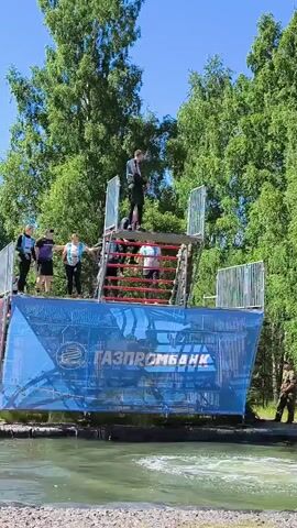 This video shows an outdoor obstacle course. In the background, there are many tall green trees. The main focus is a large blue structure with the word "ГАЗПРОМБАНК" printed on it in white. Above this structure is a metal platform with stairs. Several people are visible on the platform. One person at the very top is looking down. To the left, four people stand on the platform, three of whom are wearing dark tops and one in a light blue top. Two people are on the stairs, and another person is on the platform above them, wearing a white and pink top. In the foreground, there is a body of water. A person in a black top and pants is jumping from the platform into the water, creating a splash. Another person in a military-style uniform is standing to the right of the blue structure, looking towards the water.