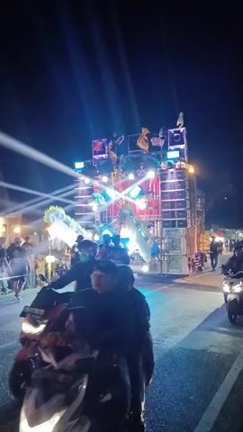 The video shows a nighttime outdoor event with a large sound system and light show. Several people are visible on and around the sound system, some appearing to be operating equipment or performing. Others are standing or walking in the foreground and midground, with some riding motorcycles. The environment appears to be a street or open area, with buildings and lights visible in the background. The overall atmosphere suggests a lively celebration or performance.