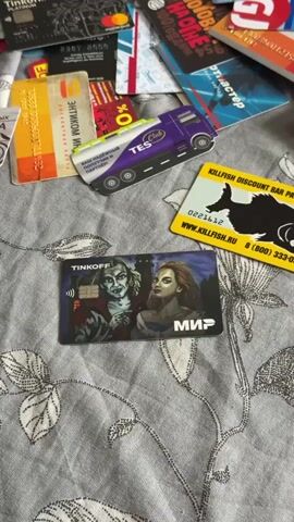 The video shows a close-up view of a collection of various cards scattered on a gray fabric surface with a leaf pattern. The cards are a mix of bank cards, discount cards, and loyalty cards. Some notable cards include a Tinkoff bank card with a vampire and woman design, a Killfish discount bar payment card with a fish graphic, and a TES Club card shaped like a truck. Other cards visible have logos and names of various companies and brands, including Yandex Money, Sportmaster, and Krasnoe & Beloe. The cards are positioned haphazardly, overlapping each other on the fabric. The camera slowly pans across the scene to display the full assortment of cards.