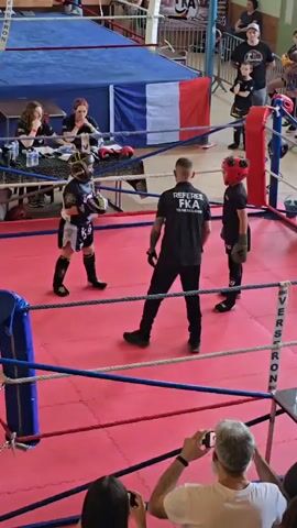 The video shows a martial arts competition taking place in a boxing ring, possibly kickboxing, featuring two young contestants wearing protective gear. A referee dressed in black attire with 'REFEREE FKA TEAMFELGELER' printed on the back monitors the match closely. The participants are engaged in sparring, throwing punches and kicks.

Surrounding the ring are spectators, including a person taking photos and two seated judges at a table, likely scoring the competition. The boxing ring is colorful, with blue and red ropes and a red canvas. A French flag is visible in the background, above the judge's table, suggesting the location. The environment seems to be an indoor gymnasium or event space adapted for martial arts competitions.