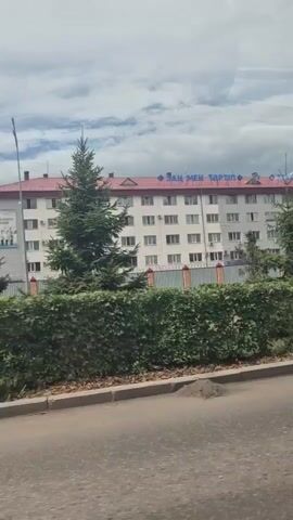 The video shows a white building with a red roof. On top of the building, there is text in blue that appears to be in a foreign language. In front of the building, there are trees and hedges. A road is visible in the foreground, and a white truck is driving on it. There is also a pedestrian crossing with red and white stripes. The sky is cloudy.