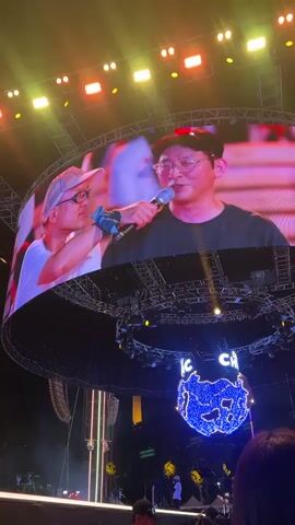 The video begins with a close-up of a large screen displaying a man wearing glasses and a black t-shirt, holding a microphone. To his left, another person in a white t-shirt and a light-colored cap is visible, also holding the microphone, their hand near the man's mouth. The man with the microphone is speaking and appears to be smiling. The screen is curved and illuminated by stage lights in red, yellow, and green. Below the screen, a large circular stage structure is visible with numerous lights and what appears to be a blue, sparkling decoration in the center, possibly representing a globe or a stylized animal, with text that seems to read "IC CH." Further down, a crowd of people is gathered, facing the stage. The overall environment is a nighttime outdoor event, likely a concert or festival, with a focus on a performance on a large stage.