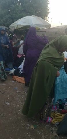 The vertical video captures a bustling outdoor market scene, likely during the daytime under soft lighting conditions. The ground is dry, reddish-brown earth. Numerous people are present, indicating a crowded environment.

At the beginning (0:00-0:01), the camera shows a wide view of people. In the foreground, a person draped in a purple garment is partially visible, while another person in blue lies or sits very low to the ground. Clothes and fabric items are piled on the ground to the right. White market canopies are visible in the background on the left.

The camera then pans and slightly zooms (0:01-0:02), focusing on a young boy with light skin, wearing a pink t-shirt with the number "11" on it and blue jeans. He walks towards the camera and looks directly at it. Behind him, more individuals in various attire, including a person in a red head covering, are seen amidst the market activity. Another person in a red and white checkered shirt and green pants walks away in the lower right.

From 0:02-0:03, the camera continues to pan, highlighting a person with dark skin wearing a purple garment with white zigzag patterns over a gray long-sleeved top. This individual is walking and carrying a black bag along with several brightly colored textile items draped over their arm, including green and blue striped fabric and a multi-colored patterned fabric. In the background, market stalls with various plastic goods like cups and bowls are visible under white canopies.

The video concludes (0:03-0:05) with a sustained shot of a man with dark skin, wearing a light cream or off-white long-sleeved traditional garment that features intricate embroidery around the neck and down the front. He also wears a small, reddish-brown patterned cap. The man is smiling broadly and looking directly at the camera, conveying a friendly demeanor. The background remains blurred with ongoing market activity.