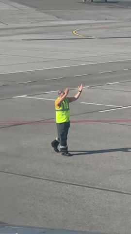 A person wearing a high-visibility vest and work clothes walks across an asphalt surface, possibly an airport tarmac. They have their hands raised in front of their face for a moment and then lower them as they walk. A yellow service vehicle is visible on the right side of the frame.