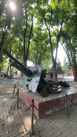 A stationary outdoor shot features an antique green anti-aircraft gun positioned on a red pedestal with a metal chain fence surrounding it. The gun is weathered and shows signs of wear. The background is filled with lush green trees, casting shadows on the ground. Sunlight beams through the trees, creating lens flare. In the distance, a statue or monument is visible behind the trees. The ground is paved with brick-like stones.
