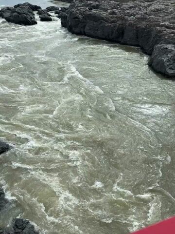The video shows a river with murky, grey-green water, rapidly flowing between jagged, dark rocks on both sides. The water is turbulent, with white foam appearing as it moves swiftly. The rocks are rough and uneven, forming the riverbanks. The overall lighting suggests a bright, overcast day. The camera angle is high, providing a direct overhead view of the river's movement.