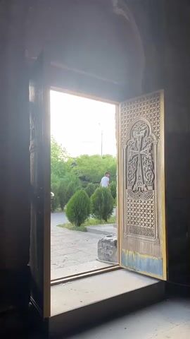 The video shows a pair of ornate wooden doors, one slightly ajar, leading outside to a courtyard area. The doors are intricately carved with religious imagery. Through the opening, lush green trees and bushes are visible, along with a paved walkway and some stone elements. The sky outside is bright, suggesting daylight. A person in the distance is partially visible. The interior of the space appears to be dimly lit and made of stone, possibly a church or monastery. The camera remains static throughout the short clip.