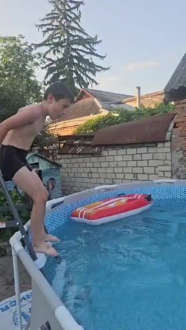 A young boy in black swim trunks stands on the edge of an inflatable swimming pool, preparing to jump. He is leaning forward, arms outstretched. An inflatable raft with red and white stripes floats in the pool. The boy jumps into the water, creating a large splash. He surfaces, coughing and wiping water from his face, and then submerges again briefly before resurfacing. The background shows a brick wall with a corrugated roof and some trees. The sky is a light blue with some clouds.