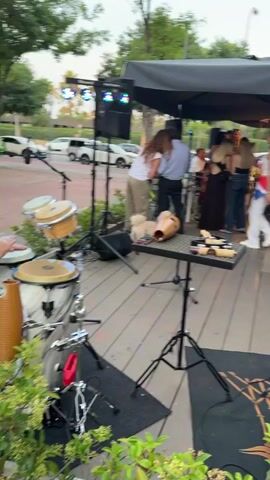 The video captures a live musical performance at an outdoor event. A band, all dressed in white, performs on a stage setup with drums and other equipment. Attendees, casually dressed, can be seen seated at tables enjoying food and drinks, while others stand around socializing. The setting is an outdoor space, likely a patio or deck area, with tables, chairs, and umbrellas providing shade. In the background, buildings can be seen, along with Cuban flags.