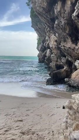 The video shows a rocky cliff face on a beach. The ocean waves are crashing against the shore. The sky is cloudy. The sand is light brown. There are some rocks on the beach.