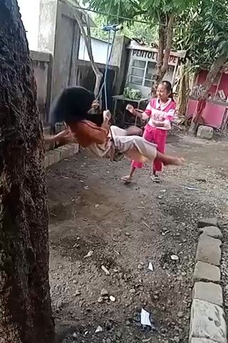 Two young females are playing on a swing outdoors. One female, wearing a hijab and a long dress, is seated on the swing, while the other female, in a pink and white striped shirt and pink pants, is pushing her. They are both smiling and laughing. The swing is suspended from a metal bar supported by trees. The ground is dirt and gravel, with some scattered debris. In the background, there is a wall, a building with a sign that reads "PENTOL POPE", and some trees. The overall atmosphere appears joyful and playful.