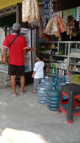 A man wearing a red shirt and black shorts stands with his back to the camera, looking into an open refrigerator. A boy in a white shirt and grey pants stands next to him, also looking into the refrigerator. The refrigerator is filled with food items. Behind them, shelves are stocked with various products, including snacks and bottled drinks. To the right of the store entrance, several large blue water jugs are stacked. A red and black plastic stool is placed on top of some of the water jugs. The ground is concrete. The scene appears to be an outdoor convenience store or small market. A woman in a brown dress is visible inside the store, lying down.
