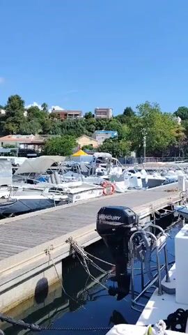 This video captures a bright, sunny day at a marina, likely in Pollica, Italy, as indicated by the context. Numerous boats, predominantly white, are docked along wooden piers. The sky is a clear blue with a few scattered white clouds.

In the foreground, a sturdy wooden dock is visible, extending into the mid-ground. Several powerboats and smaller vessels are moored to it. One boat, seen from the stern, has "Deborah" clearly written on its transom, which is covered by a blue canvas. Another boat's outboard engine prominently displays the brand "Mercury." Electrical cables and hoses are coiled on the dock near the boats.

In the middle of the video, a person wearing a red shirt and with curly brown hair is seen from behind, moving along the dock and appearing to handle some equipment near a boat. The camera briefly pans to the right, following the person's movement and revealing more of the marina.

The background features green, tree-covered hills dotted with various buildings. A distinctive, light-colored stone tower or historic structure is visible atop one of the hills, adding a local architectural element to the scene. The overall impression is a vibrant and active marina environment on a beautiful day.