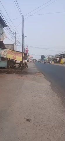 The video shows a street scene with several motorcycles passing by. On the left side of the street, there are some buildings and what appears to be a small shop. There are also utility poles and overhead wires. The street itself is paved, and there are some loose gravel and debris on the side of the road. The sky is overcast.