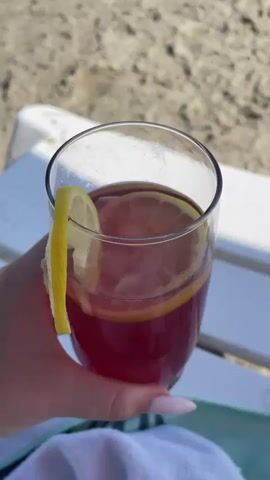 The video shows a first-person view of a person holding a glass of red liquid with a lemon slice, possibly a beverage or cocktail, on a beach. The background features a sandy beach, with a white lounge chair in the foreground. Beyond the beach, the ocean stretches out to the horizon under a clear blue sky, suggesting a sunny day. The waves are gently rolling onto the shore. In the distance, a person can be seen near the water. The scene conveys a sense of relaxation and enjoyment, likely during a vacation or a leisurely day at the beach. The time of day appears to be midday or early afternoon, given the bright sunlight and clear sky. The location is identified as Skala Avdiron, Greece. There is no visible text in the media file.