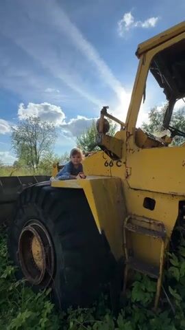 A young child is positioned on top of the front loader of a yellow construction vehicle. The child is wearing a blue shirt and is facing the camera. A shoe and sunglasses are on the surface near the child. The construction vehicle appears weathered and possibly abandoned, with the sun shining through the frame. The background consists of trees and a blue sky with white clouds. The video appears to be a short, static shot.