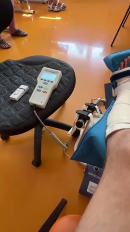 The video shows a first-person view of a person's leg connected to a medical device. The device appears to be a continuous passive motion (CPM) machine, used for physical therapy after surgery or injury. The leg is secured in a brace with blue padding. A control unit for the CPM machine is visible on a nearby stool. The scene is an indoor environment, likely a clinic or physical therapy room, with an orange floor and medical equipment in the background. There are other people in the background, partially visible, who appear to be in a similar setting.