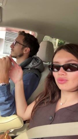 Inside a car, a man wearing glasses, a blue shirt, and a gray vest is driving. A woman with long brown hair, sunglasses, and a brown top is in the passenger seat, her seatbelt fastened. She is looking towards the camera and occasionally turning her head toward the driver. She touches the driver's hand in a gesture of affection. The car is moving along a street with buildings visible through the windows. The interior of the car is light colored. The car has headrests covered in patterned material. There is a brown bag visible near the woman.