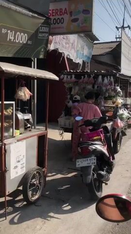 The video shows a street scene with several food vendors. In the foreground on the left is a food cart selling what appears to be fried snacks displayed in a glass case. The cart has a price sign indicating RP 14.000. Next to it, a motorcycle is parked. In the background, a person in a pink outfit is standing behind a stall selling various items in plastic bags. Further down the street, more stalls with goods displayed are visible. The environment appears to be a sunny day on a street with buildings and power lines overhead.