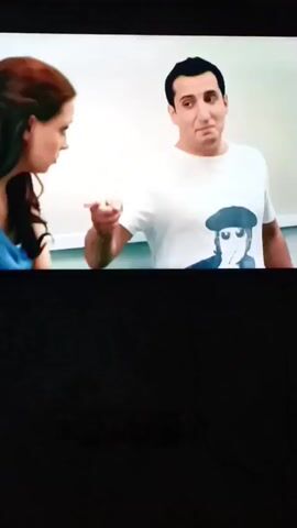 A woman with long brown hair tied back and a blue tank top smiles as a man in a white t-shirt with a graphic print points at her. He is speaking to her, and she appears to be responding. The background is a plain wall with some light-colored paneling. The image is a screenshot from a video, and the bottom portion of the screen is black.