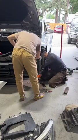 Two individuals are working on the front of a car in what appears to be a garage or workshop. The car's hood is open, revealing the engine compartment. The front bumper has been removed and is lying on the floor in front of the vehicle. One person, wearing a beige t-shirt and tan pants, is standing and appears to be working on the upper part of the car's front end. The other person, wearing a dark t-shirt and brown pants, is kneeling and working on the lower part of the car's front end, near the wheel well. Tools and car parts are scattered on the concrete floor around them. The environment is cluttered with shelves, equipment, and other vehicles visible in the background.