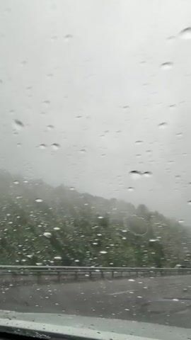 The video shows a view from inside a car, looking out through the windshield on a rainy, foggy day. Raindrops are visible on the glass, and the windshield wiper moves periodically to clear the view. Outside, there's a road with a guardrail, and a hillside covered in green vegetation is visible through the fog. The sky is overcast and gray, contributing to the overall gloomy atmosphere.