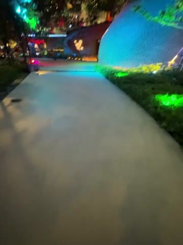 The video opens on a pathway at night, illuminated by various colored lights. To the right is a large, circular, mesh-like structure covered in green plants and lit with blue and green lights. To the left, a metal structure holds a series of bright blue spotlights. In the background, a building with a unique, undulating facade is lit with green and blue lights. Trees and greenery are scattered throughout the scene. As the video progresses, the lighting on the building shifts from green to blue and the overall color scheme intensifies. A few people are visible walking in the distance near the spotlights. The pathway itself is smooth and appears to be made of concrete. The scene suggests an outdoor public space, possibly a park or plaza, decorated for an event or display.