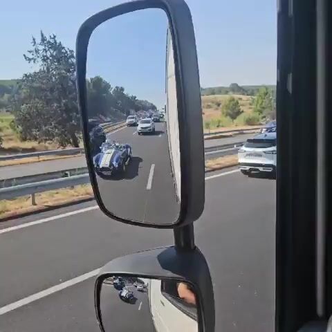 The video captures a view from the driver's side mirror of a large vehicle, likely a truck, driving on a highway. The mirror reflects a clear blue sky and a sunny day. In the main part of the mirror, a vintage blue and white sports car, possibly a Shelby Cobra, is seen driving in the same direction as the viewer. Several other vehicles, including white vans and cars, are also visible on the highway ahead of the sports car. The highway has multiple lanes and is bordered by a guardrail and dry, grassy hills with scattered trees. A smaller, lower mirror below the main one reflects the road behind the viewer's vehicle, showing other cars. The camera is stationary within the frame, showing the movement of the highway and the vehicles through the mirrors.