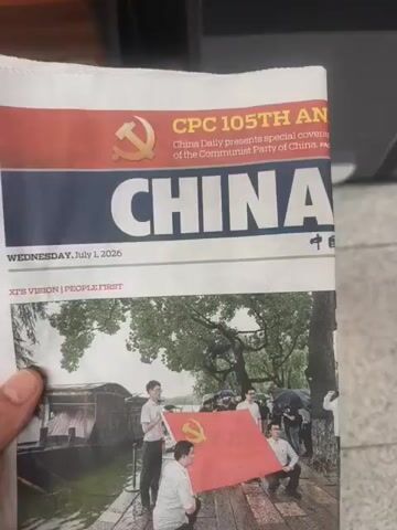 A person's hand holds a copy of the China Daily newspaper. The newspaper's headline reads "CPC 105TH AN..." and "CHINA". Below that, the date is Wednesday, July 1, 2026. A picture on the newspaper shows several people holding a large red flag with a yellow hammer and sickle symbol on it. The text below the image says "Putting people first at". The person is standing in front of a dark-colored bin with labels that read "RESTAFFALD" and "OTHER WASTE". The hand then moves the newspaper towards the opening of the bin. The video ends as the newspaper is about to be placed in the bin.