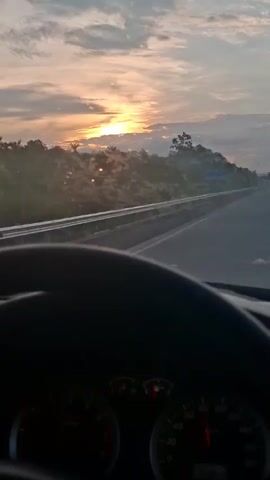 The video shows a first-person perspective from inside a car. The driver's steering wheel is in the foreground, and the dashboard with gauges is visible below it. The car is on a highway, and the view through the windshield shows a scenic landscape. The sun is setting or rising, casting a warm glow over the sky, which is filled with clouds. The road stretches ahead, with trees and vegetation on the sides. A guardrail separates the highway from the adjacent land. A sign is visible on the left side of the road, displaying text and numbers, possibly indicating a toll or distance marker. The overall impression is that of a peaceful drive through a natural environment during a beautiful sky phenomenon.