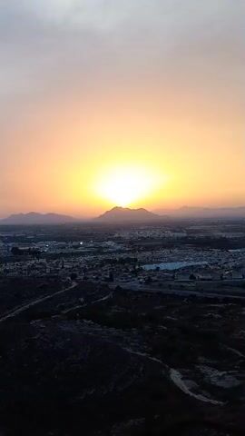 This is a time-lapse video showing a sunset over a residential area and distant mountains. The camera is positioned on a hillside overlooking the scene.

The subjects include the sky, which transitions from a bright, clear yellow-orange near the horizon to a pale, hazy orange and then a muted, dark orange at the top of the frame. A prominent mountain range forms the backdrop, silhouetted against the setting sun. Below the mountains lies a sprawling urban area, characterized by rows of houses with red-tiled roofs and interspersed with roads and commercial buildings. In the foreground, a dark, rugged hillside with sparse vegetation is visible.

The activity captured is the natural event of a sunset. The sun, a brilliant orb, descends behind the central mountain peak, casting a warm glow across the sky and illuminating the landscape below. As the sun sinks lower, the light intensifies in its immediate vicinity, creating a halo effect, and the colors of the sky deepen. The urban lights begin to appear as the daylight fades.

Notable details include the time of day, which is clearly sunset. The weather appears clear, with a slight haze contributing to the softened colors of the sky. The location cues, such as the urban layout with red-tiled roofs and the arid landscape, suggest a Mediterranean or similar climate. The overall emotion evoked is one of tranquility and the serene beauty of nature. There is no visible text in the video.
