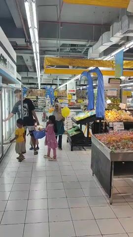 A family of four is shopping in a brightly lit supermarket. On the left, a young boy in a yellow shirt stands by a freezer. Next to him, a taller individual, possibly an older sibling or parent, wearing a black t-shirt and shorts, is also looking into the freezer. A young girl in a pink dress is standing in the middle of the aisle, holding a blue basket. A woman wearing a hijab and a teal and grey outfit is walking towards the right side of the frame, holding a yellow balloon. Various products are visible on shelves and displays throughout the store, including produce and candy. The floor is tiled.